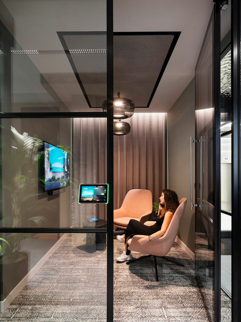 Person sitting in a modern, glass-walled office room with a mounted screen and tablet. Room has two pink chairs, a patterned carpet, and overhead lighting.