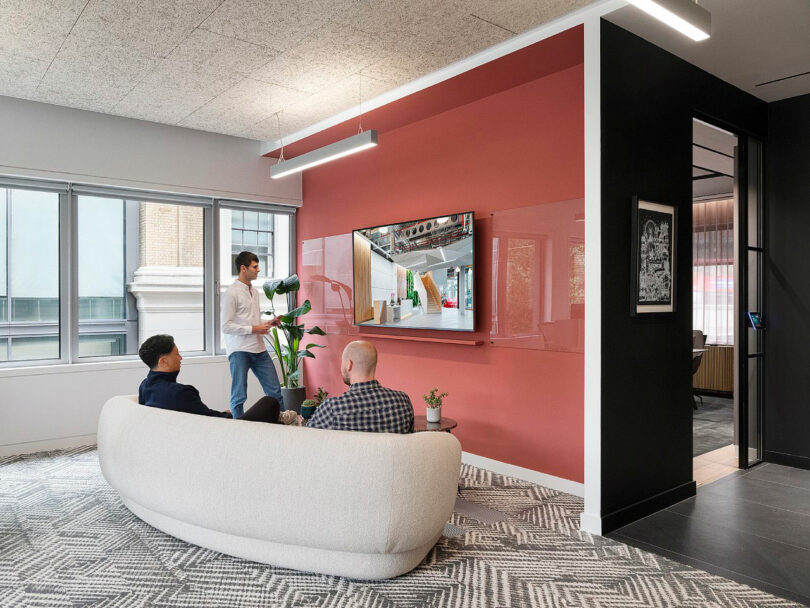 A person gives a presentation on a wall-mounted screen to two seated individuals in a modern office space with large windows and a textured carpet.