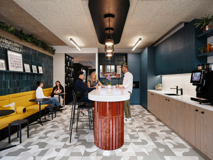 Five people are socializing in a modern kitchen area with a central island, tiled floor, yellow seating, and decorative plants and lights.