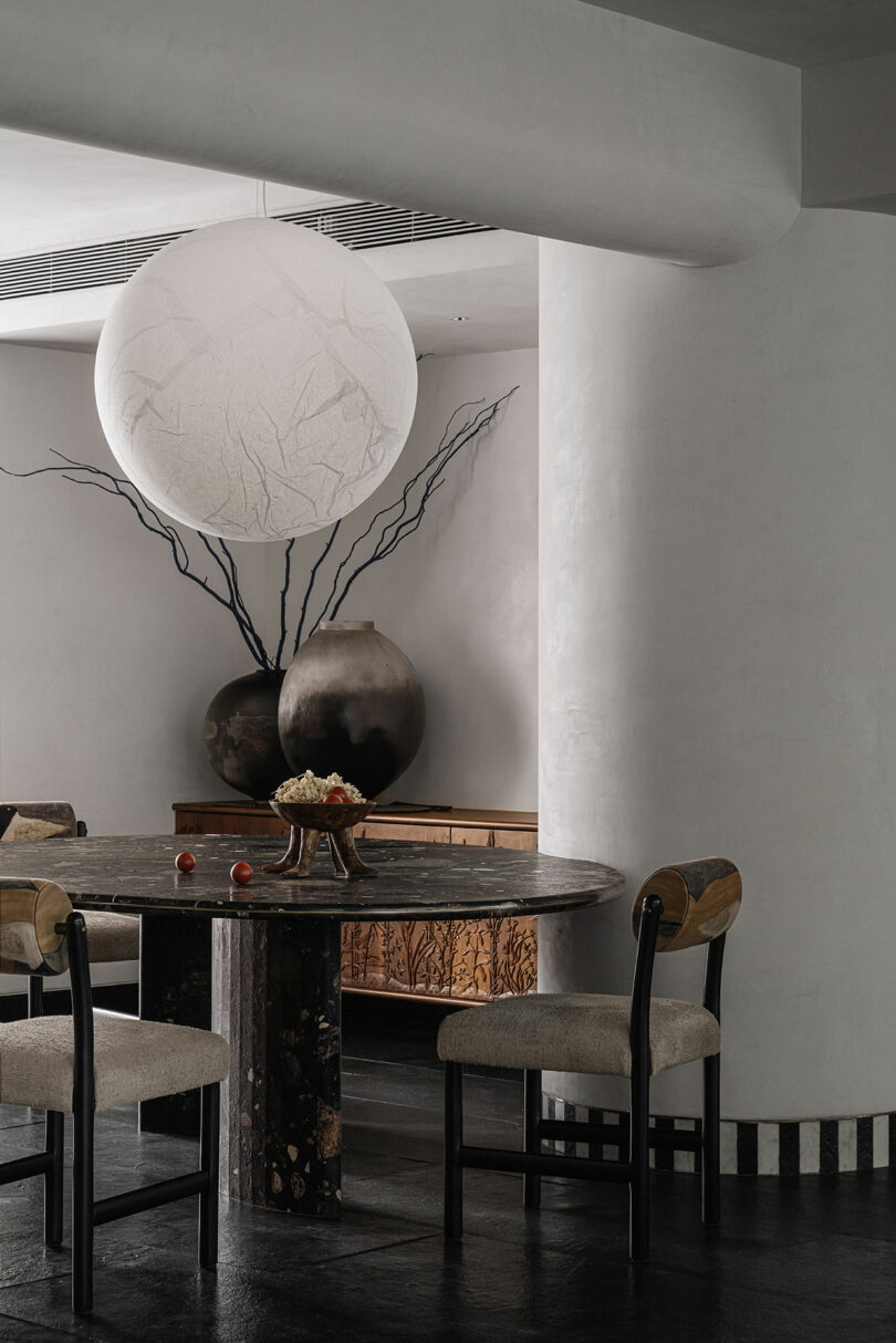 A modern dining room by Studio Nishita Kamdar features a round table, sleek black chairs, and a large spherical lamp. Decorative vases adorn the console against pristine white walls, complemented by a sophisticated dark floor.