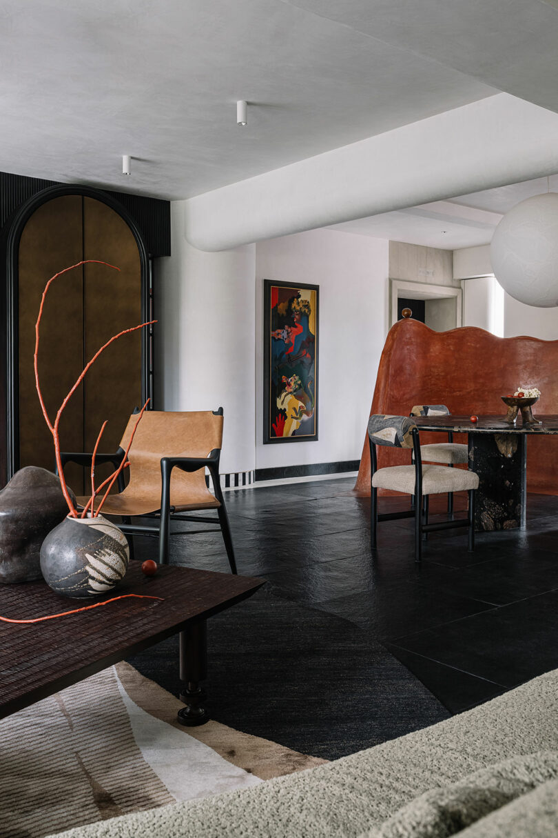 Modern interior by Studio Nishita Kamdar featuring a leather chair, large pottery vase with red twigs, abstract artwork on the wall, and a curved partition in a spacious room with dark flooring and minimalist decor.