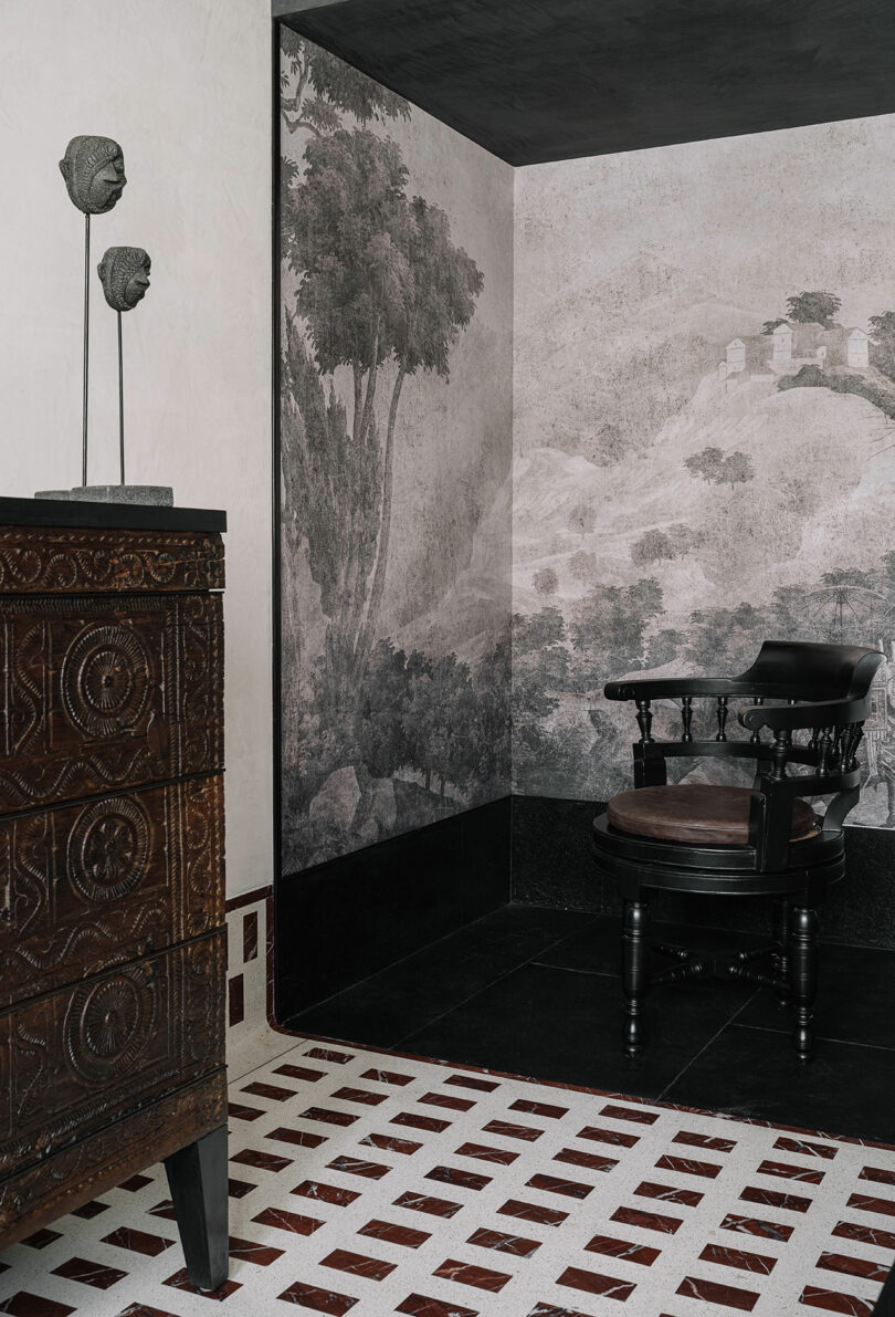 A corner room designed by Studio Nishita Kamdar features a wooden chair, a chest of drawers, and wall art depicting a landscape. Two sculpted heads are elegantly displayed on a stand atop the chest, complemented by the floor's patterned design.