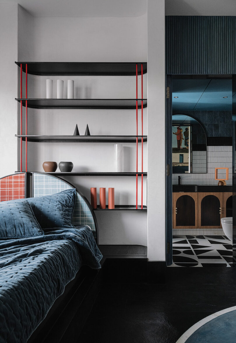 Modern bedroom by Studio Nishita Kamdar features a blue bed and red-accented shelves displaying minimal decor, offering a view into a bathroom with geometric tiles and wooden cabinetry.