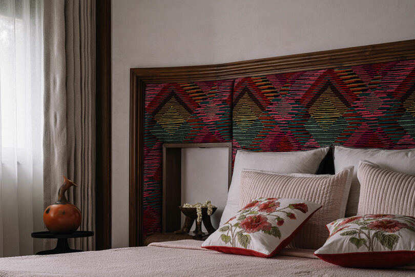 A cozy bedroom designed by Studio Nishita Kamdar features a colorful geometric pattern headboard, floral-patterned pillows, and a side table with a ceramic vase. Light filters through sheer curtains, adding a touch of serene elegance.