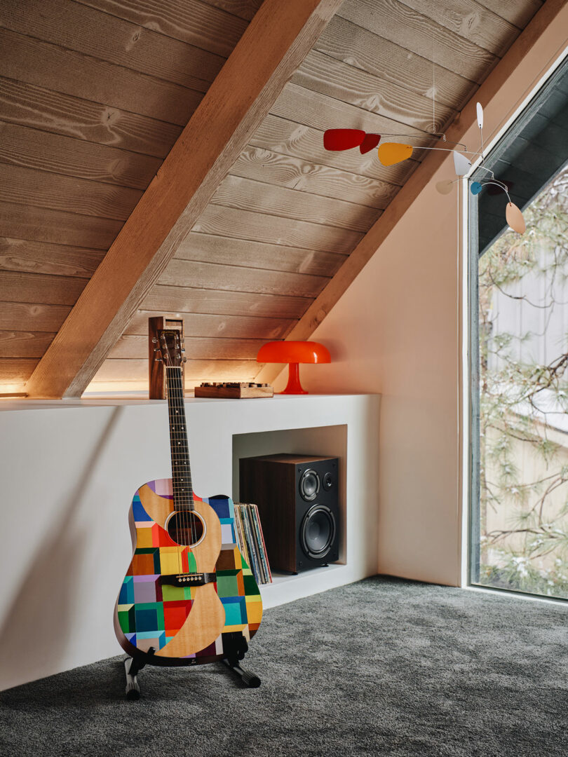 A colorful acoustic guitar on a stand in a cozy attic room, with a lamp, speaker, and books nearby. A modern mobile hangs from the ceiling near a window.