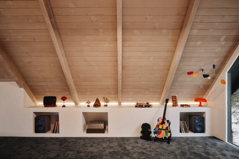 A cozy attic room with slanted wooden ceiling, featuring shelves with books, a colorful guitar, speakers, and decorative items.