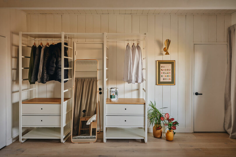 A modern, organized closet with white shelving, hanging clothes, a full-length mirror, plants, and wall art on a beige paneled wall.