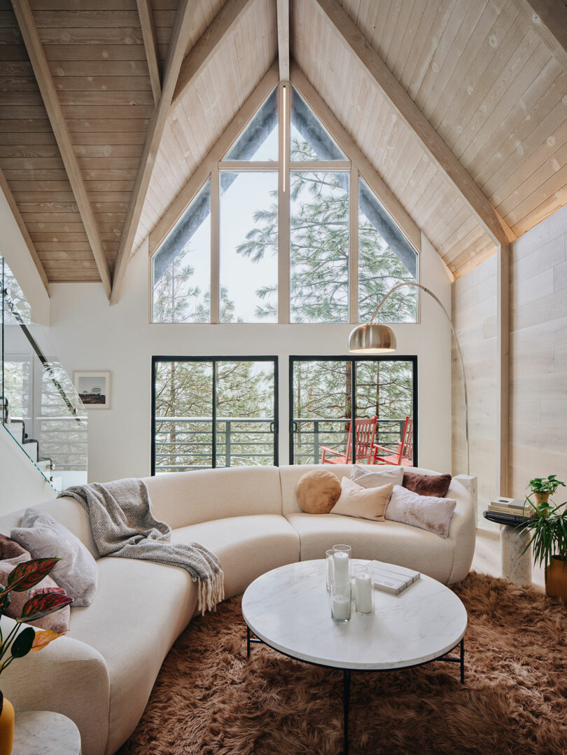 A cozy living room with a vaulted ceiling, white curved sofa, marble coffee table, floor lamp, and large windows showcasing a scenic outdoor view.