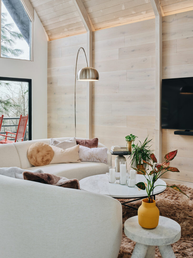 A cozy living room with a beige sectional sofa, round marble coffee table, and a floor lamp. Decor includes throw pillows, a fur rug, candles, and houseplants. Large windows provide natural light.