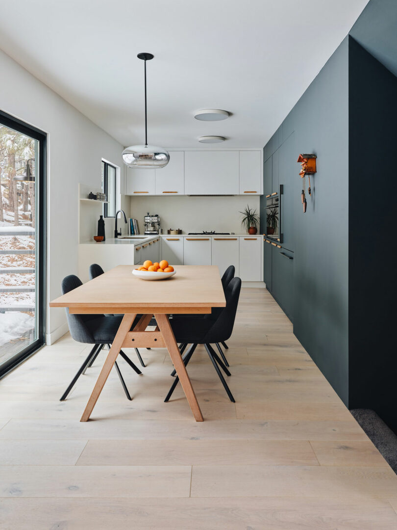 Modern kitchen with a wooden dining table, black chairs, and a bowl of oranges. White cabinets and dark accent wall in the background. Large window on the left.