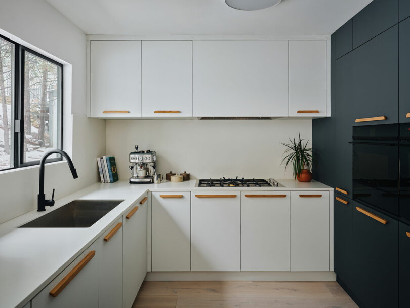 Modern kitchen with white cabinets, a black sink, a gas stove, and a coffee maker on the counter. A plant and cookbooks are displayed near the window with outdoor views.