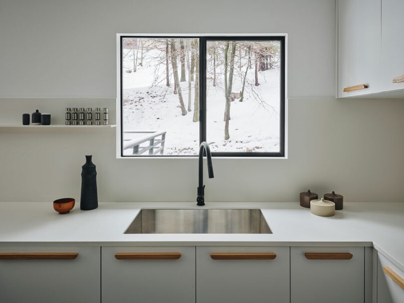 Modern kitchen with white cabinets and minimalist decor, featuring a large window view of a snow-covered landscape with trees.