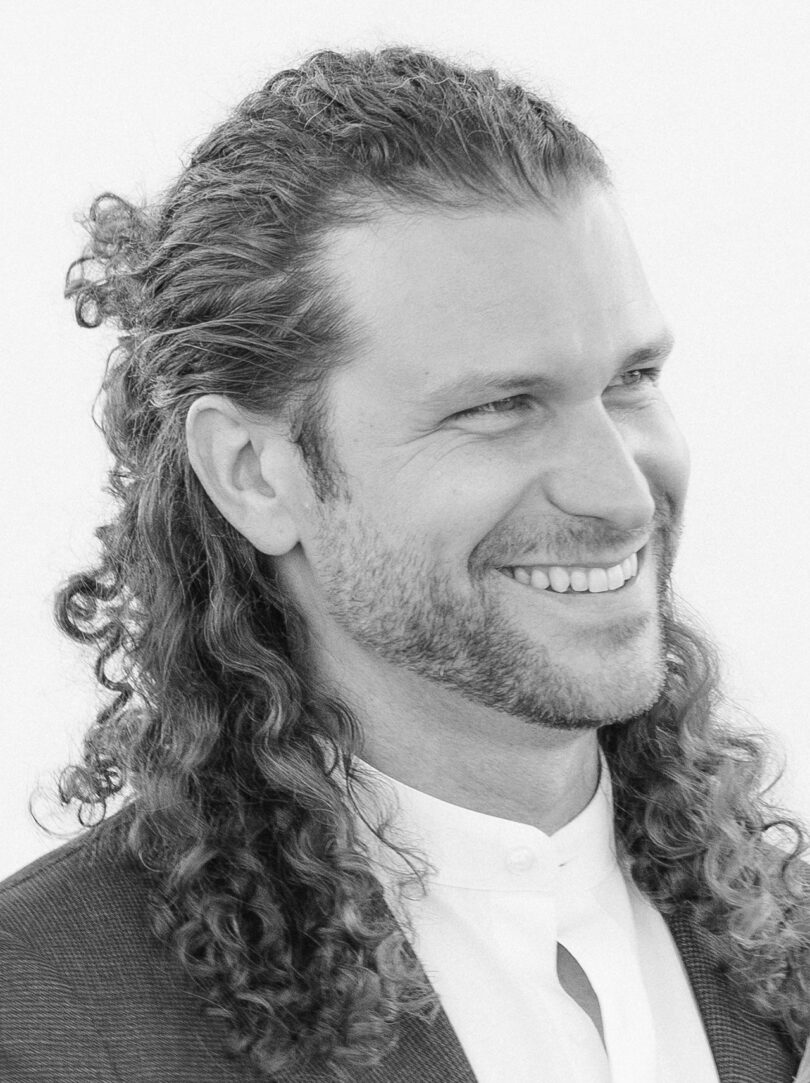 A man with long, curly hair smiles while looking to the side. He is wearing a suit jacket over a button-up shirt.