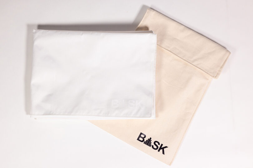 Tela blanca doblada junto a un bolso BASK beige con un logo sobre una superficie blanca.