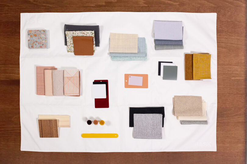 A variety of fabric and material samples are artfully arranged on a white surface, including swatches in neutral, earthy, and pastel tones, complemented by paint chips and small round color samples.