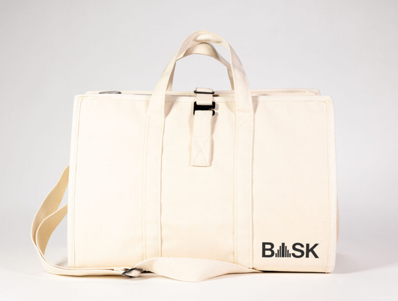 El bolso BASK de two-point es un accesorio de lona beige con asas resistentes y una correa para el hombro, que presenta el icónico logotipo de la colaboración BASK en un lugar destacado en la parte delantera.