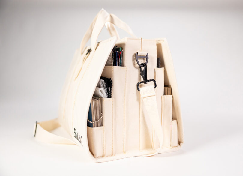 The beige BASK Bag from two-point features multiple external pockets, perfect for holding books and essentials. A sturdy shoulder strap is attached via a metal clip, while the bottom corner is subtly labeled "BAG.