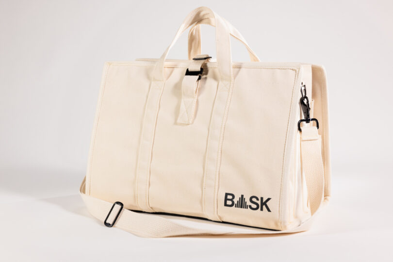 Un bolso de lona beige con asas y una correa ajustable para el hombro, con el logo. "DISFRUTAR" en el costado.