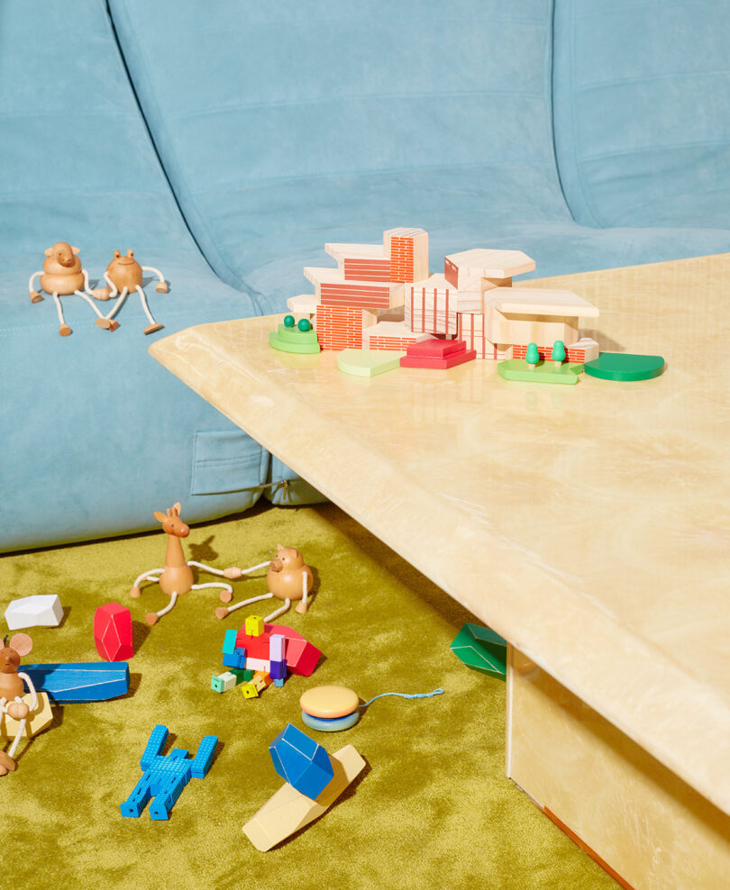 Toys are scattered on a green carpet near a beige table and blue couch, including building blocks and small figurines.