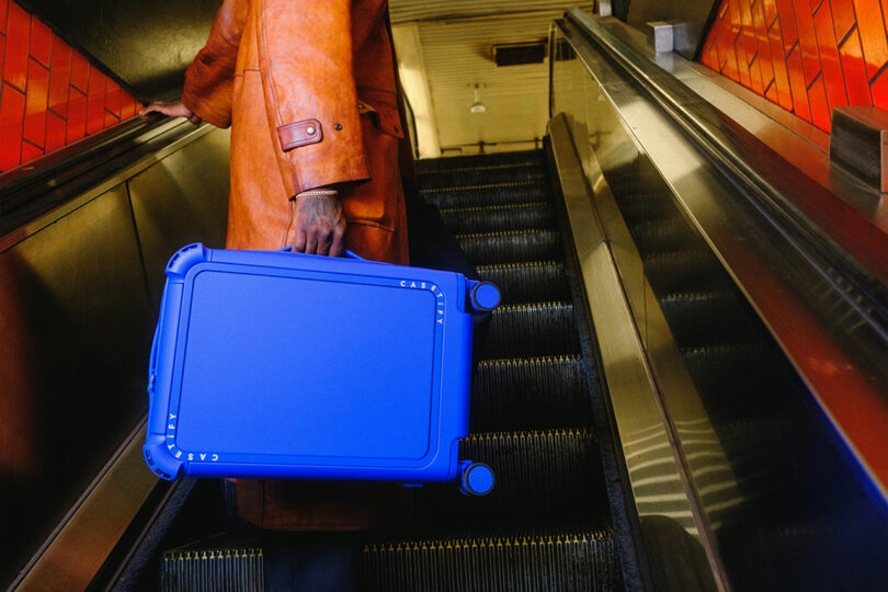 Person in an orange coat holds a blue suitcase while ascending an escalator with red tile walls.