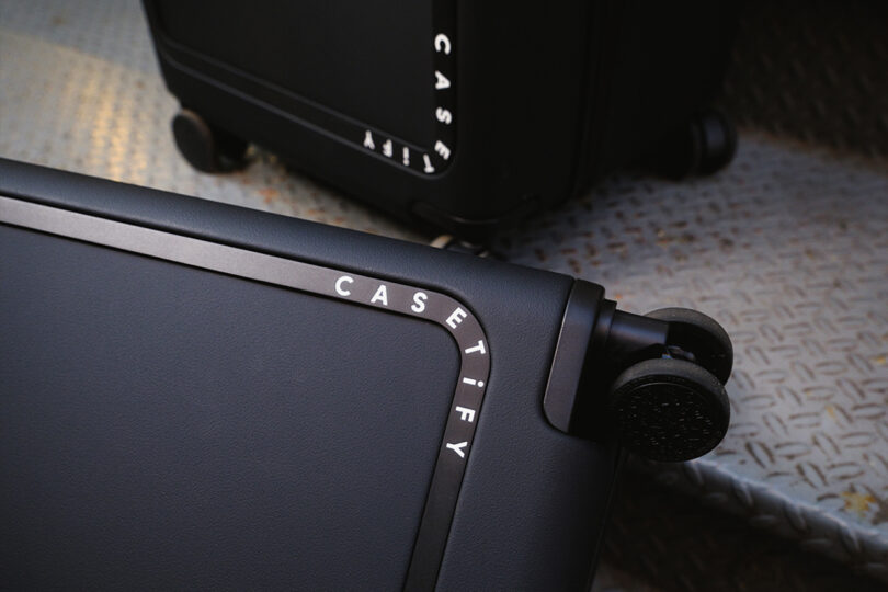 Close-up of black Casetify suitcase corners with wheels on a textured metal surface.