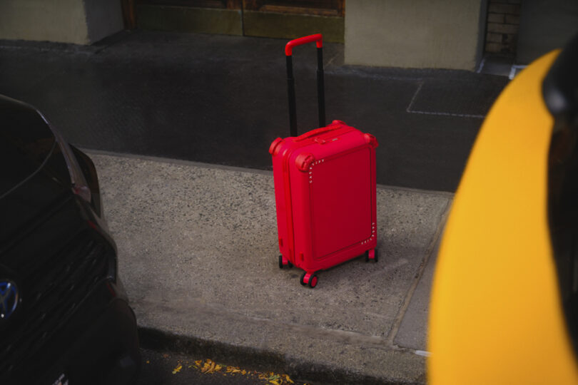 A red suitcase stands on a sidewalk between two parked vehicles.