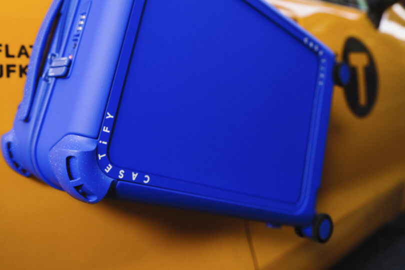 A blue suitcase is attached to the side of a yellow taxi with its wheels facing outward.