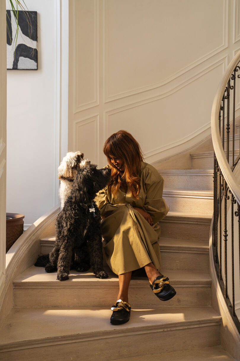 A person sits on stairs with two dogs, one black and one white. They are inside a well-lit room next to a black and white painting.