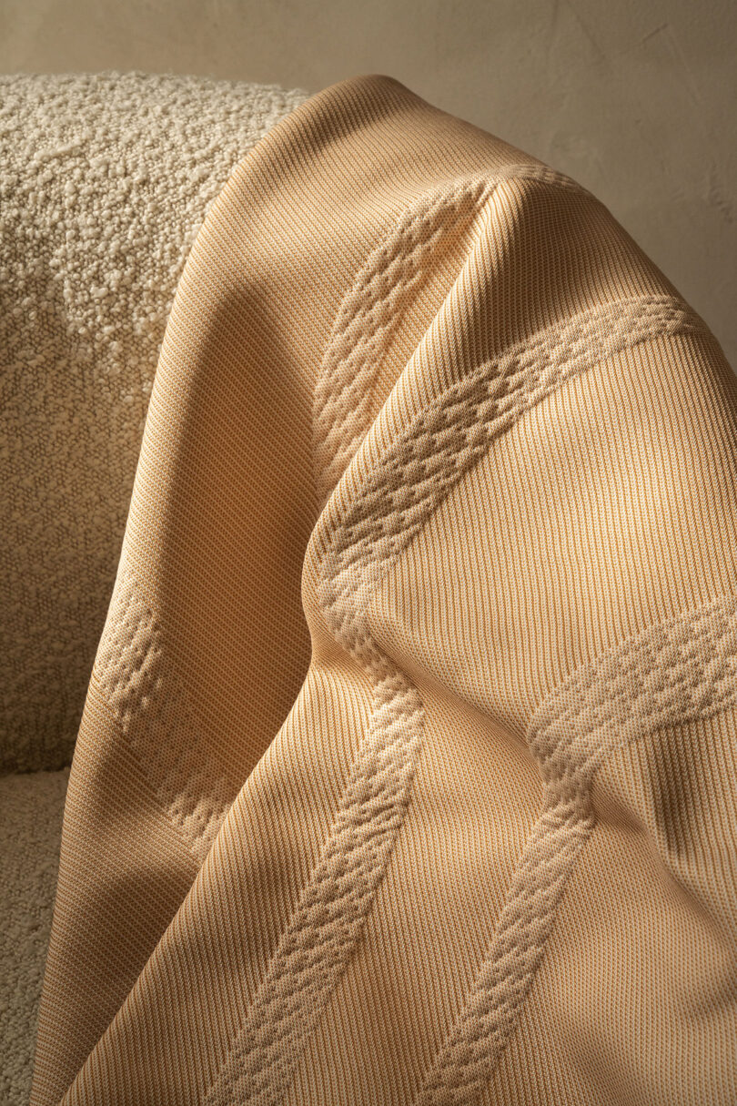 Close-up of a beige textured fabric with intricate stitching, draped over a cream-colored woven chair.