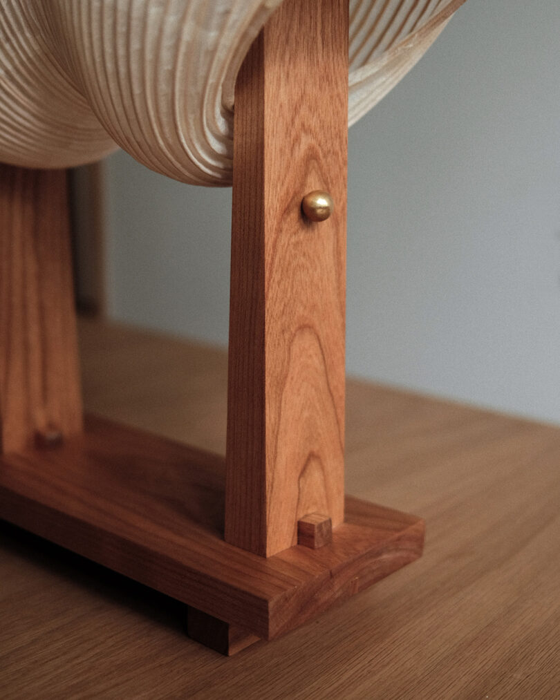 Close-up of a wooden lamp base with a textured white lampshade. The wooden stand features a brass detail and sits on a wooden surface.