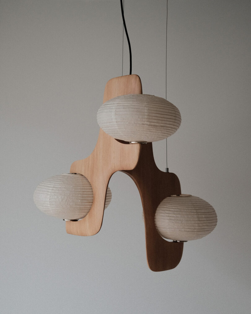 A modern wooden chandelier with three white, oval-shaped lampshades hangs from the ceiling against a plain background.