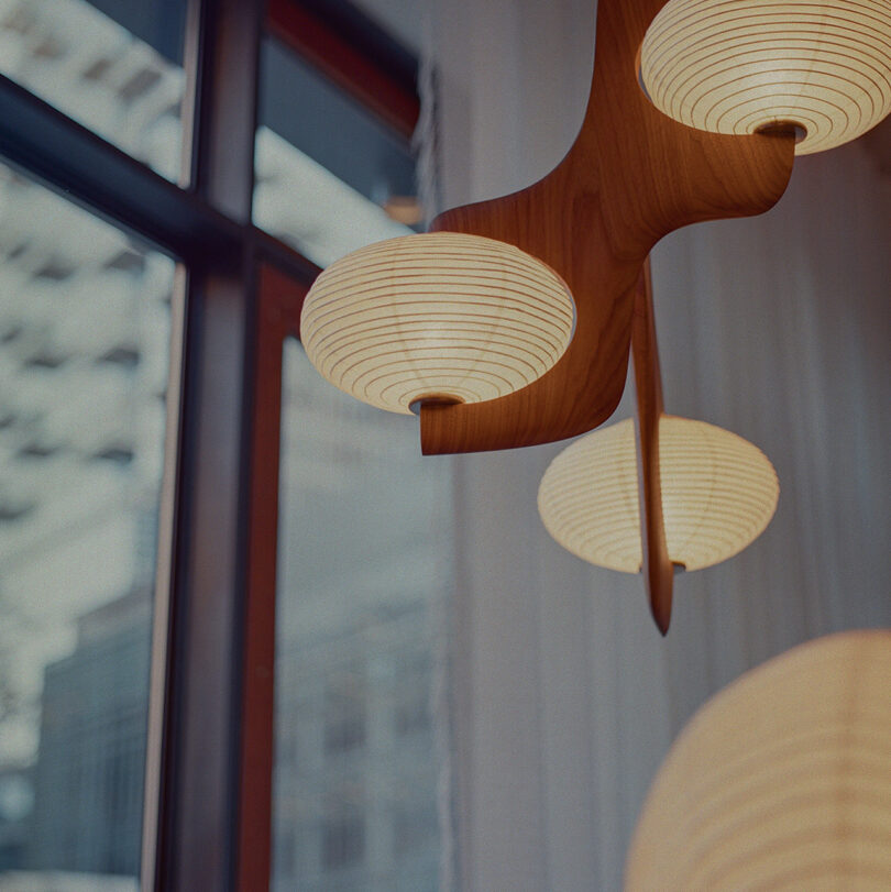 Wooden light fixture with round, ribbed paper shades hangs near a large window. Background shows blurred cityscape.