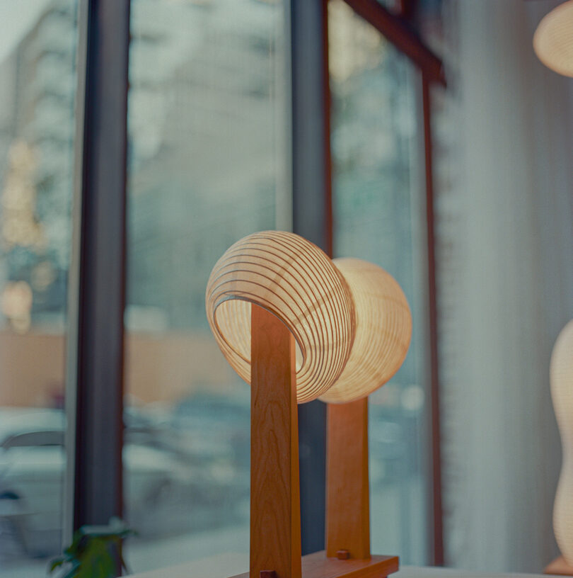 A pair of modern, spherical lamps with wooden bases are placed near a large window, allowing daylight to filter through.