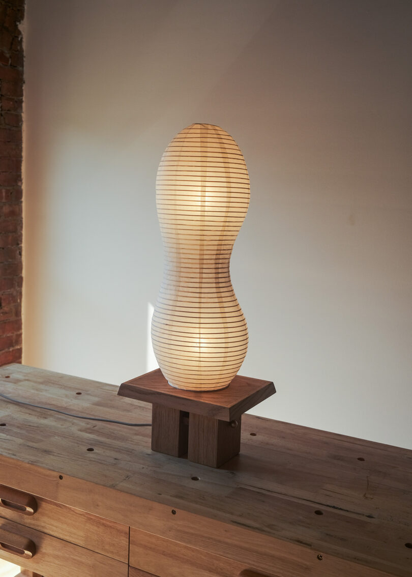 A uniquely shaped lamp with a wooden base sits on a wooden table, casting a soft glow against a plain wall.