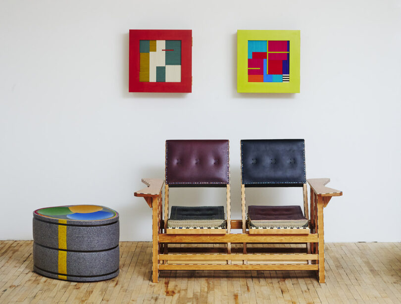 Wooden bench with black and burgundy cushions, a round ottoman with colorful top, and two abstract paintings on a white wall.