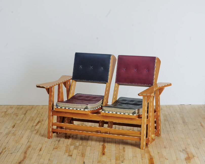 A vintage wooden double chair with a blue and a red cushioned seat, placed on a wooden floor against a plain white wall.