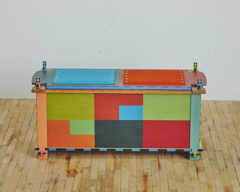 A colorful, geometric-patterned wooden bench with a patchwork design stands on a light wood floor against a plain wall.