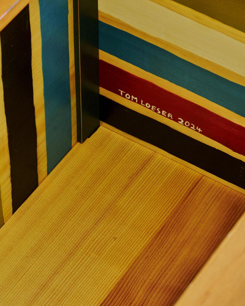 Wooden box interior with multicolored vertical stripes on the sides and a wooden base. The name "Tom Loeser 2024" is painted on one of the stripes.