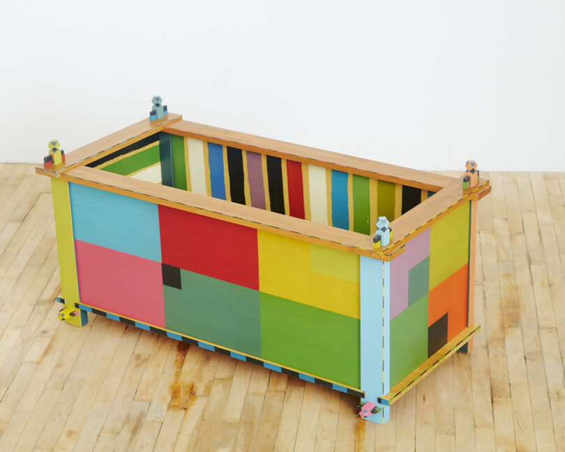 A colorful, rectangular wooden box with a pixelated design stands on a wooden floor.