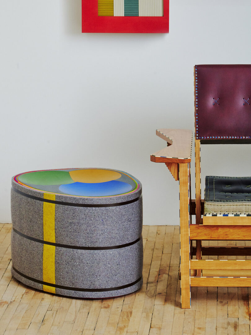 Colorful circular ottoman next to a wooden bench with a burgundy cushioned backrest. Abstract art hangs on a white wall.