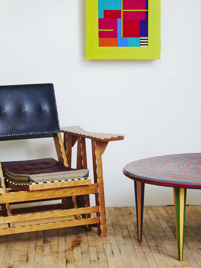 A wooden chair with a leather seat, a colorful framed abstract artwork on the wall, and a round table with a spiral pattern on a wooden floor.