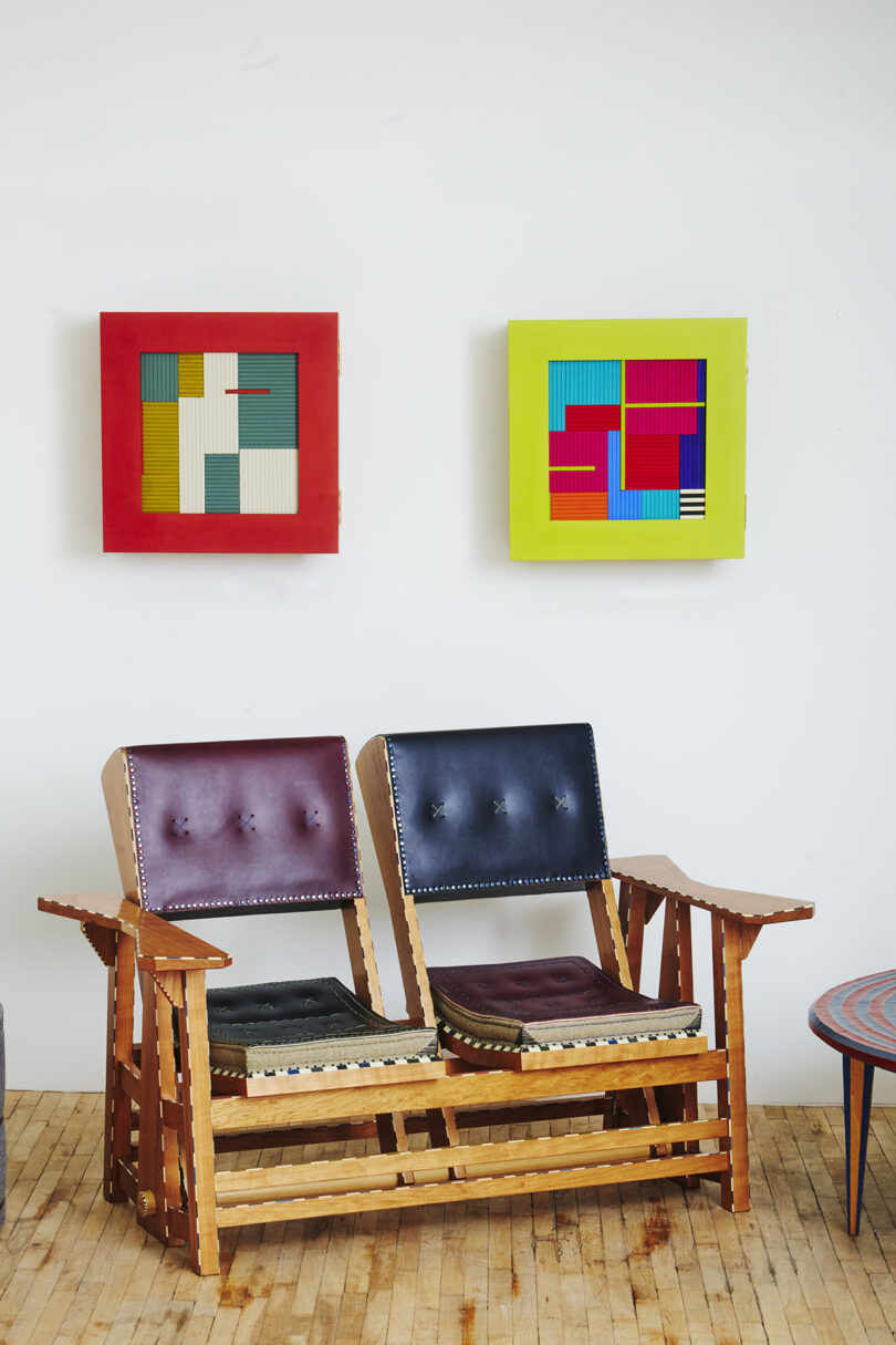 A wooden bench with two cushioned seats sits beneath two colorful abstract paintings on a wall.