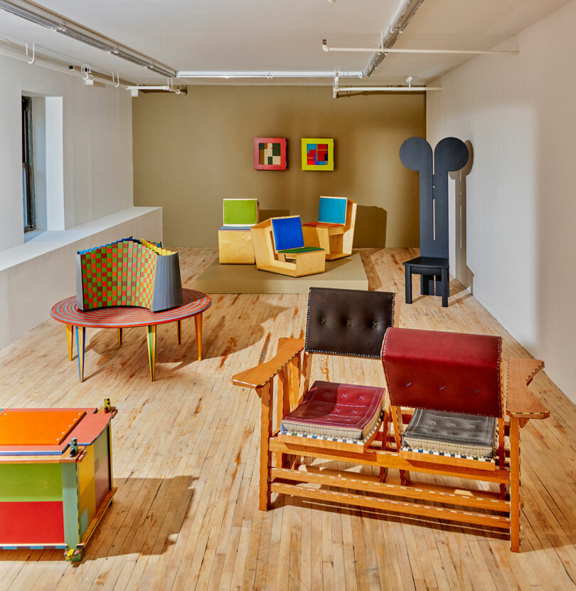 In Colorama at Superhouse, a brightly lit room showcases colorful, geometric furniture with mismatched patterns on a wooden floor. A small sculpture graces the back wall, completing the eclectic scene.