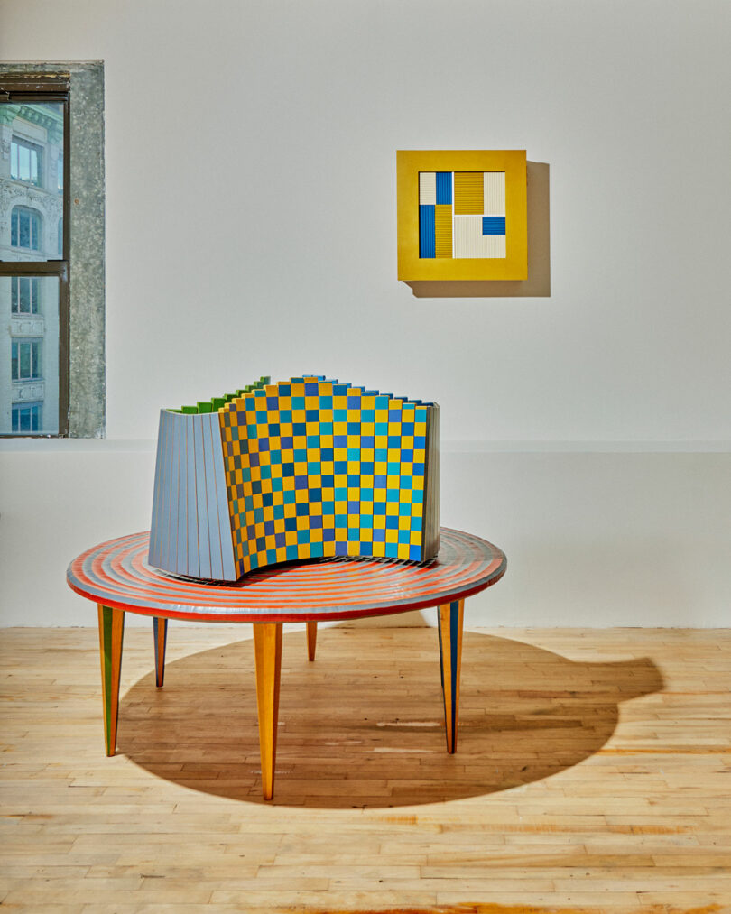 On a wooden table sits a sculpture adorned with a colorful checkered pattern, part of the Colorama at Superhouse exhibit. Nearby, a geometric artwork hangs gracefully on the wall, creating an intriguing dialogue between shapes and hues.