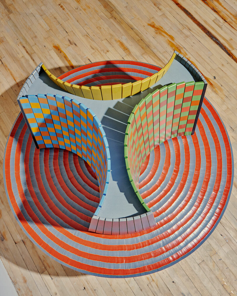 Colorama at Superhouse features a vibrant abstract sculpture with three curved walls in yellow, green, and blue standing on a red and white concentric circle floor, all set atop a wooden surface.