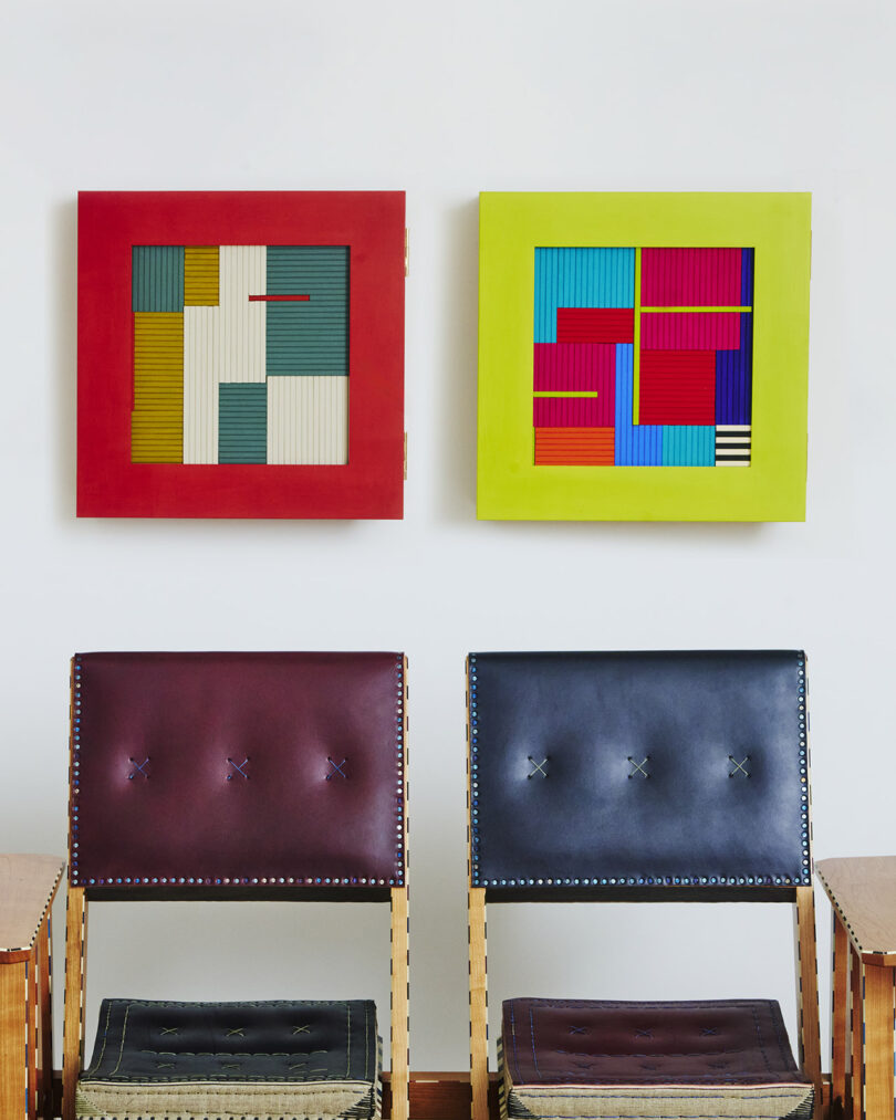 Two abstract geometric artworks hang above two leather chairs with wooden frames. The left artwork has a red frame, and the right one has a yellow frame.