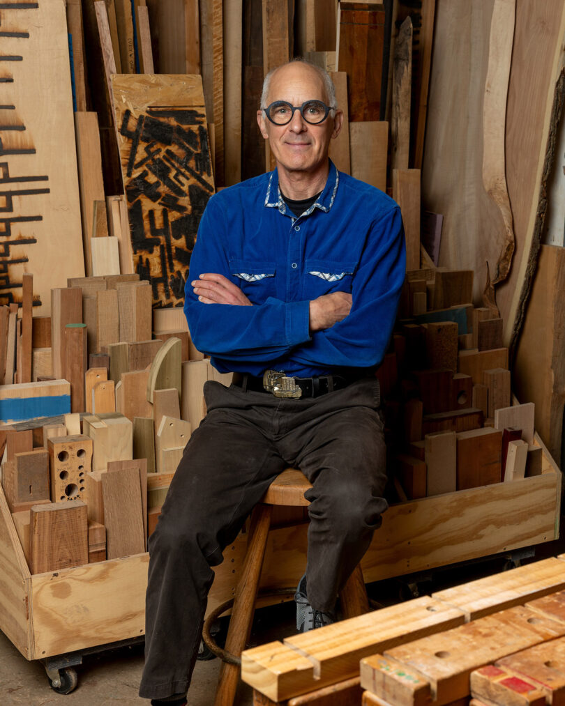 A man in a blue shirt sits on a stool in a woodshop, surrounded by wooden planks and boards.