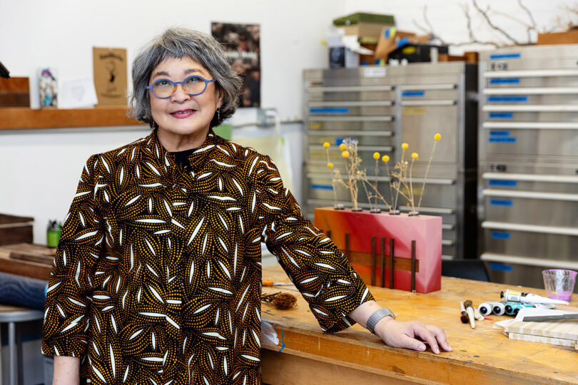 A person with gray hair and glasses stands in an art studio, wearing a patterned top. Various art supplies and a square vase with flowers are on the table.