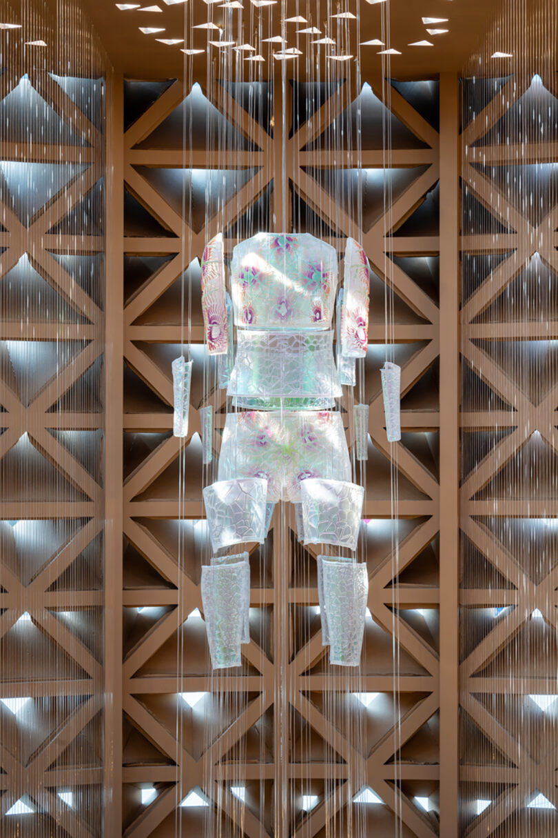 Translucent armor-like suit suspended in an ornate, geometric display area with intricate lighting effects.