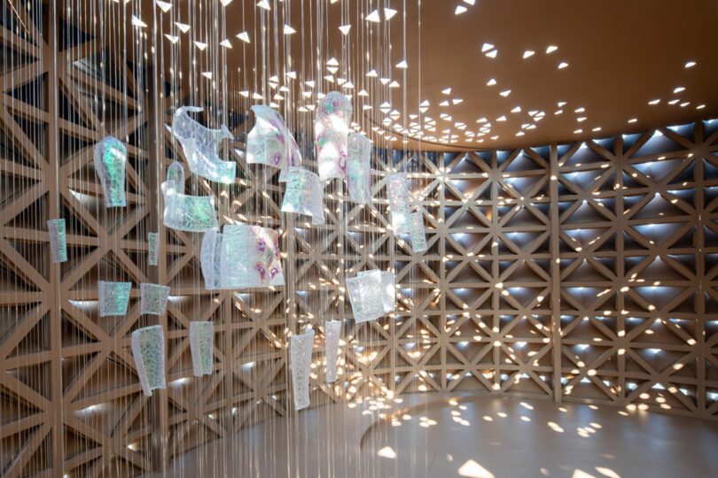 Hanging translucent art pieces on strings in a circular room with geometric wall patterns and light filtering through small openings.